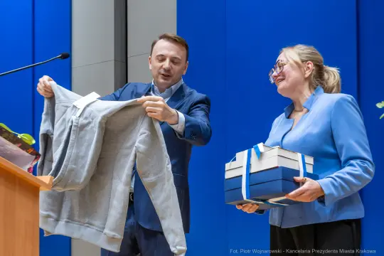 Mężczyzna wręcza kobietę szary sweter oraz niebieskie pudełka z prezentami.