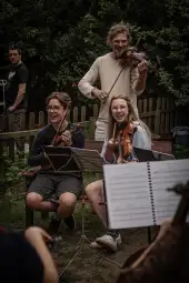 Grupa osób grających na skrzypcach w ogrodzie, radosna atmosfera.