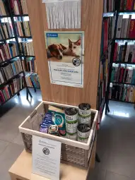 Kosz z darami dla zwierząt w bibliotece, plakat promujący akcję pomocy dla psów i kotów.