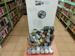 Zbiór puszek z jedzeniem w bibliotece, obok ulotki informacyjnej o akcji charytatywnej.