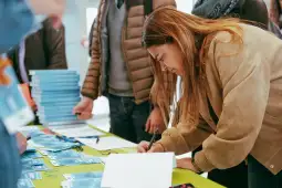 Kobieta podpisuje dokumenty przy stole z książkami i identyfikatorami.