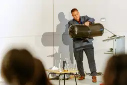 Mężczyzna w garniturze przelewa zawartość czarnego worka do naczynia na stole.