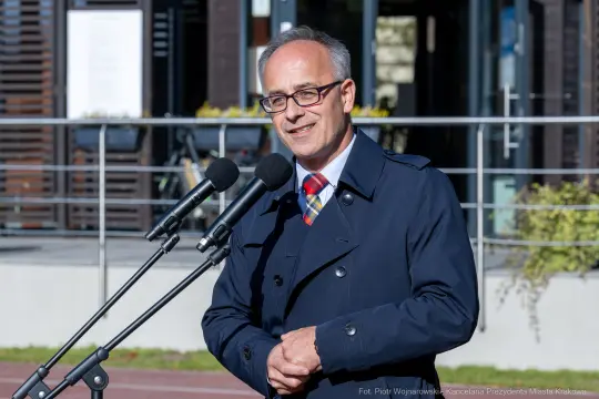 Mężczyzna w okularach i granatowym płaszczu przemawia przy mikrofonach.