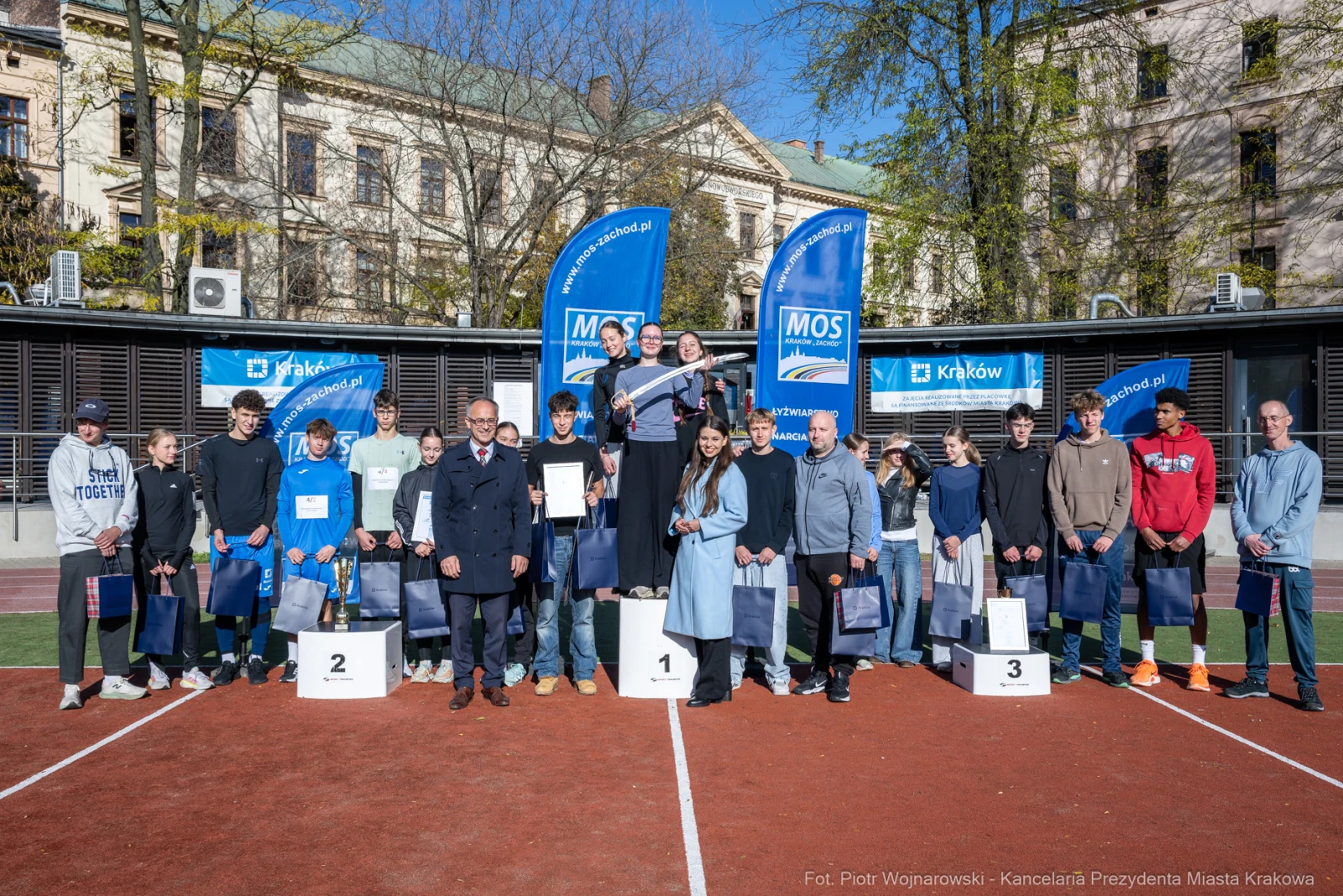 Młodzi sportowcy na podium z nagrodami, flagi i dyplomy w tle.  Autor: Piotr Wojnarowski