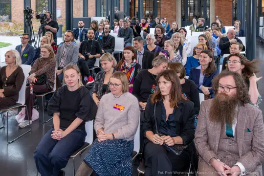 Grupa ludzi siedzących na wydarzeniu, różnorodne reakcje i styl ubioru.