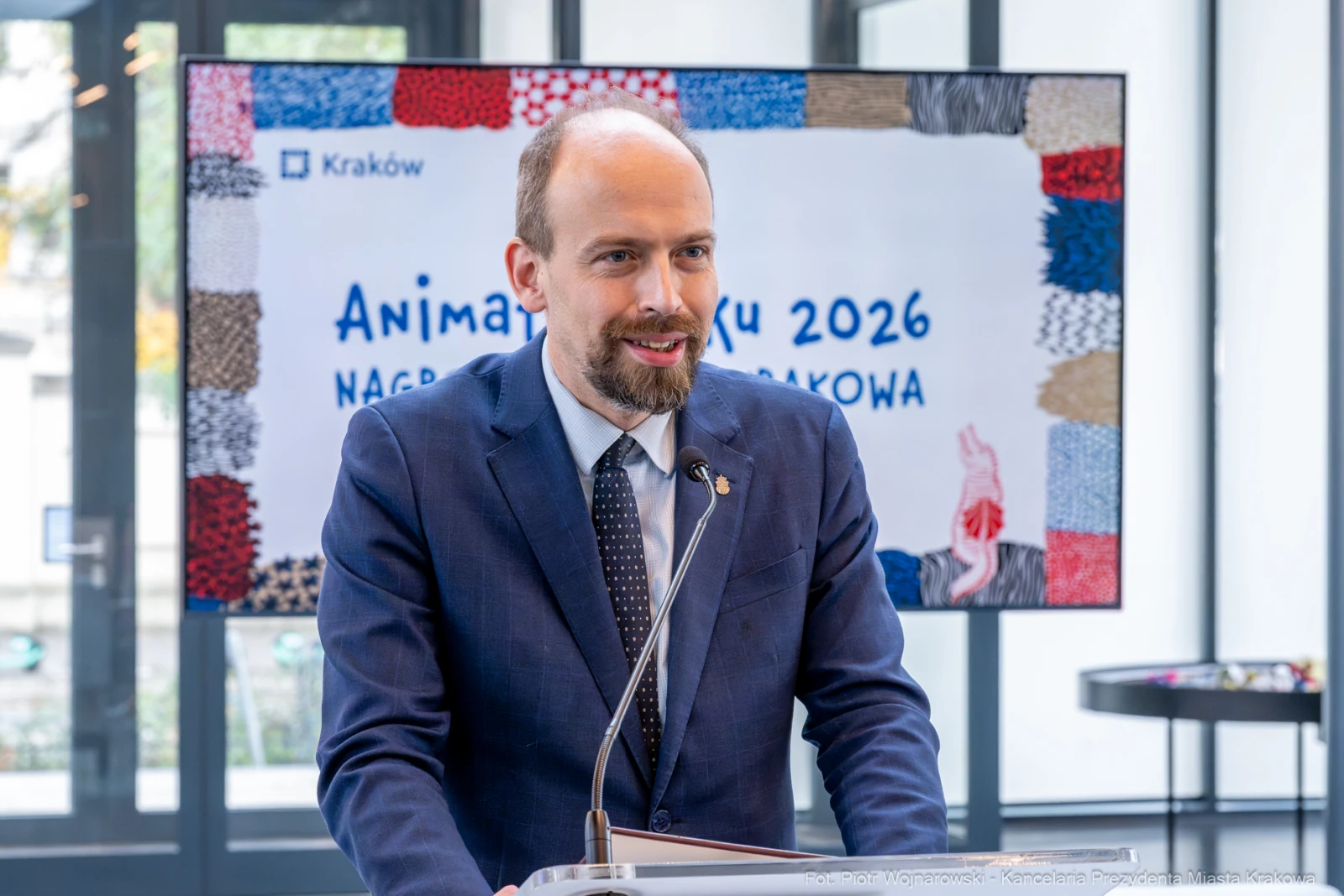 Mężczyzna w garniturze przemawia podczas wydarzenia w Krakowie. W tle widoczny plakat z napisem o nagrodach.  Autor: Piotr Wojnarowski