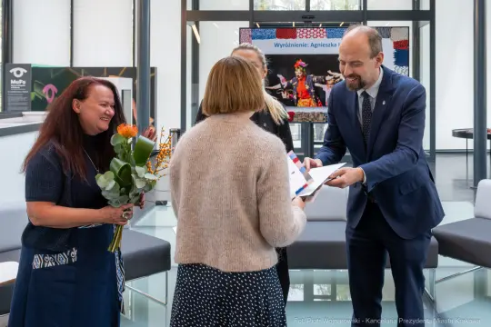 Wręczenie nagrody, uśmiechnięte osoby, kwiaty i dyplom w nowoczesnym wnętrzu.