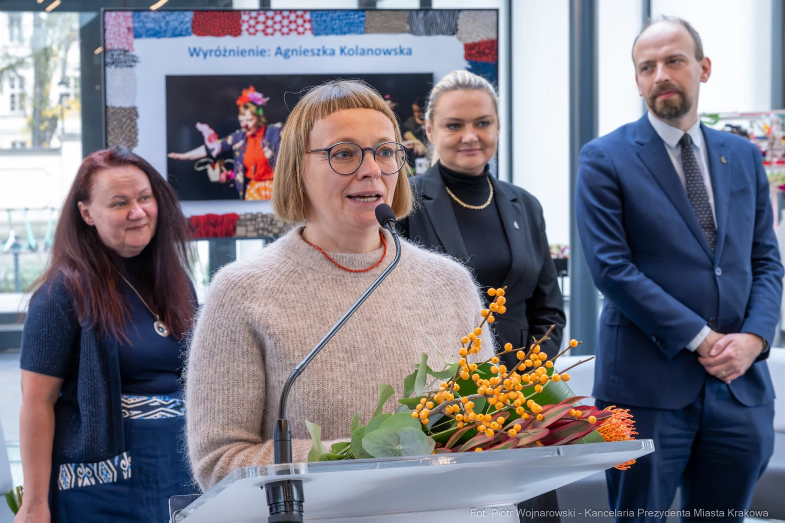 Kobieta przemawia przy mikrofonie, obok niej stoją inne osoby, w tle wystawa.  Autor: Piotr Wojnarowski