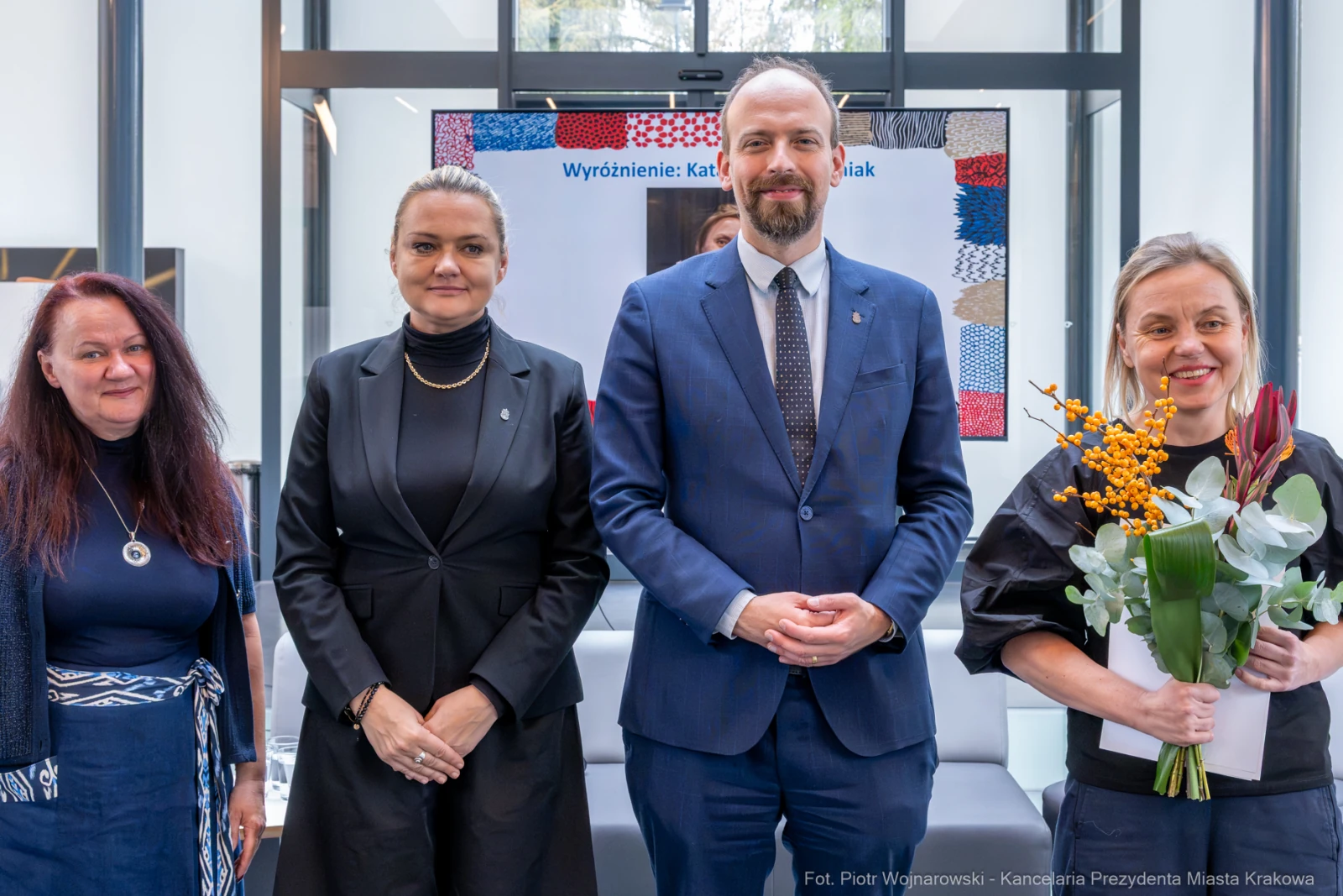 Cztery osoby w formalnych strojach, jedna z bukietem kwiatów, w nowoczesnym wnętrzu.  Autor: Piotr Wojnarowski
