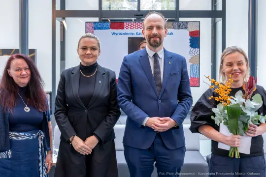 Cztery osoby w formalnych strojach, jedna z bukietem kwiatów, w nowoczesnym wnętrzu.