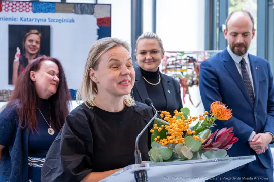 Kobieta przemawia z bukietem kwiatów, w tle inni uczestnicy wydarzenia.