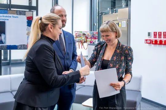 Trzy osoby w formalnych strojach, jedna wręcza dokument, w tle nowoczesne wnętrze.