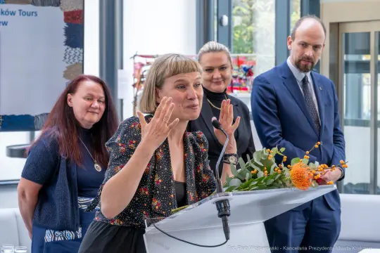 Kobieta przemawia przy mównicy, w tle inne osoby, kwiaty na stole.