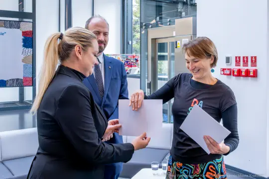 Wręczenie dokumentów podczas uroczystości. Dwie kobiety i mężczyzna w tle.