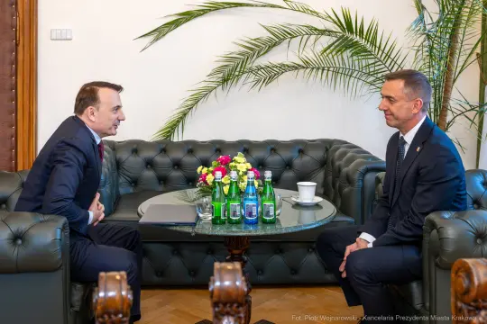 Dwóch mężczyzn rozmawia przy stole z napojami i kwiatami w eleganckim wnętrzu.