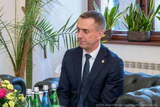 Mężczyzna w garniturze siedzi przy stole z butelkami wody i kwiatami.