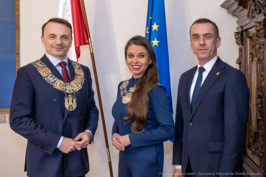 Trzech mężczyzn i kobieta w eleganckich strojach, z flagami Polski i UE w tle.