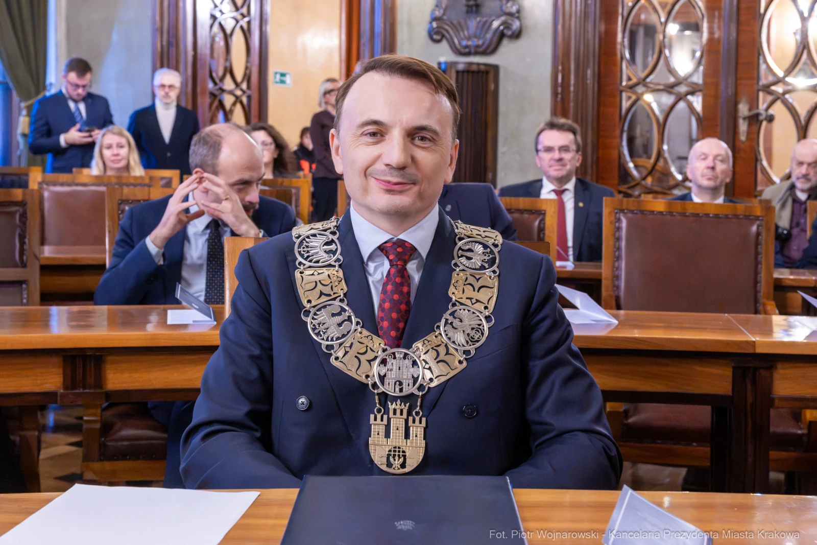 Mężczyzna w garniturze z łańcuchem na szyi, siedzący w sali obrad z innymi osobami w tle.  Autor: Piotr Wojnarowski