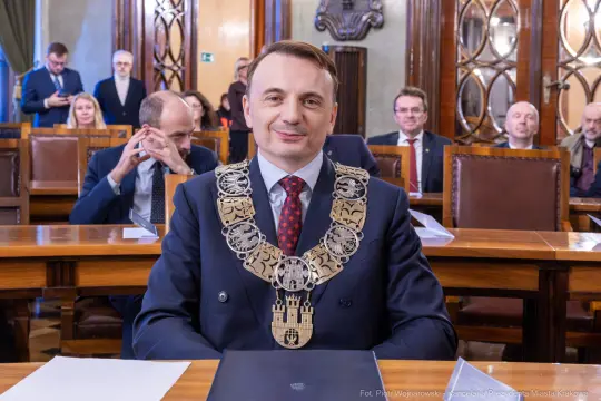 Mężczyzna w garniturze z łańcuchem na szyi, siedzący w sali obrad z innymi osobami w tle.