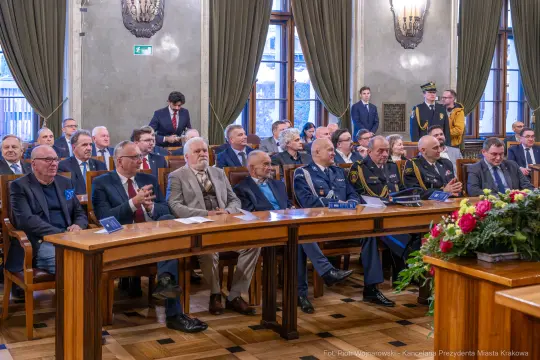 Uczestnicy uroczystości siedzący przy długim stole w sali z oknami i dekoracjami.