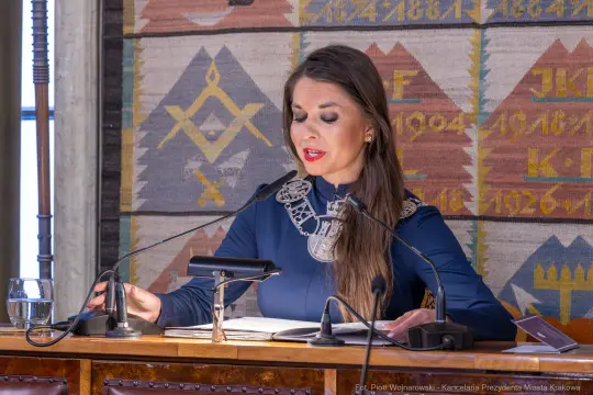 Kobieta w niebieskiej sukience z ozdobnym kołnierzem przemawia przy stole.