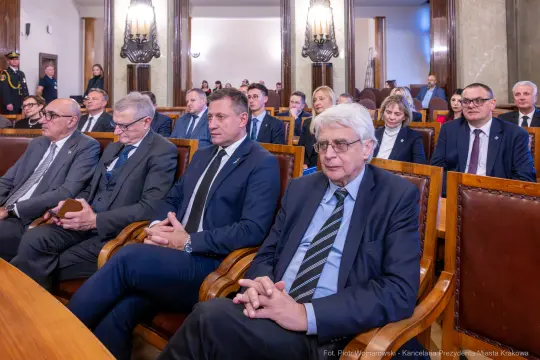 Grupa osób siedzących w sali, wśród nich mężczyźni w garniturach i kobiety w eleganckich strojach.