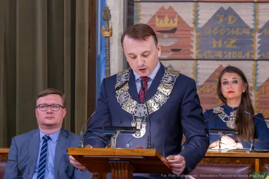 Mężczyzna w łańcuchu prezydenckim wygłasza przemówienie, w tle kobieta i mężczyzna w garniturach.