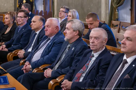 Grupa ludzi siedzących w sali, ubranych w eleganckie stroje, podczas uroczystości.