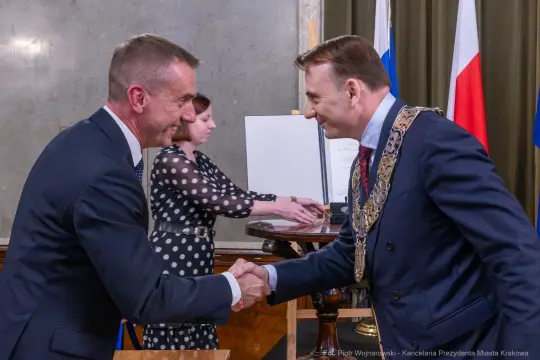 Dwaj mężczyźni uściski dłoni, jeden w łańcuchu prezydenckim, w tle flagi.