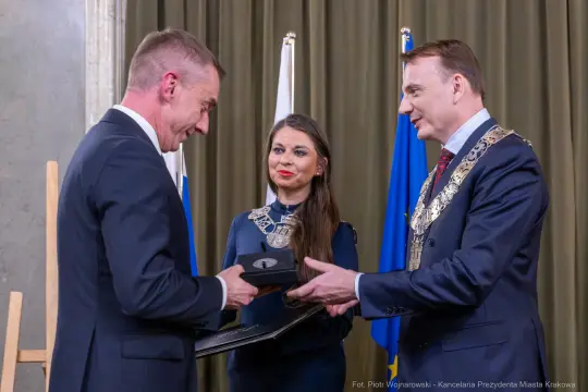 Wręczenie nagrody przez dwóch mężczyzn, kobieta obserwuje, w tle flagi.