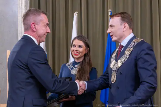 Ceremonia wręczenia nagrody z uściskiem dłoni między mężczyznami i uśmiechającą się kobietą w tle.