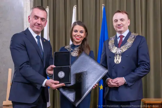 Trzy osoby w eleganckich strojach, trzymające medal i dokument w ceremonialnym otoczeniu.