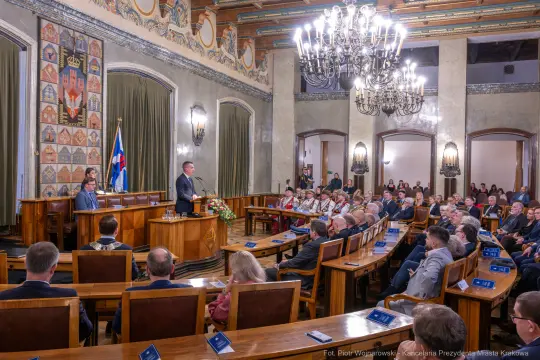 Sesja w sali obrad z mówcą i słuchaczami, dekoracje na ścianach, flagi i kwiaty.