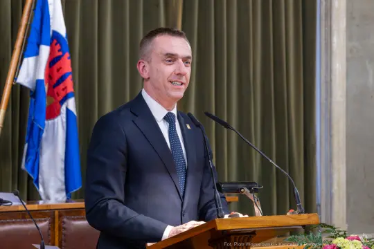 Mężczyzna w garniturze przemawia z mównicy, w tle flagi i zasłona.