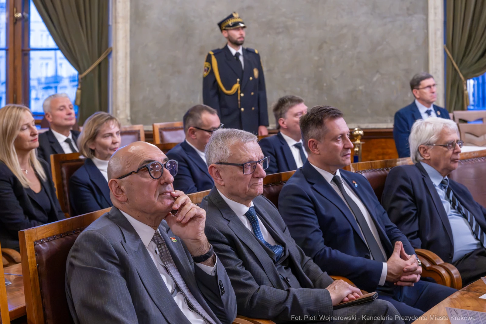 Grupa ludzi w formalnych strojach siedzących w sali obrad, z jednym strażnikiem w mundurze.  Autor: Piotr Wojnarowski
