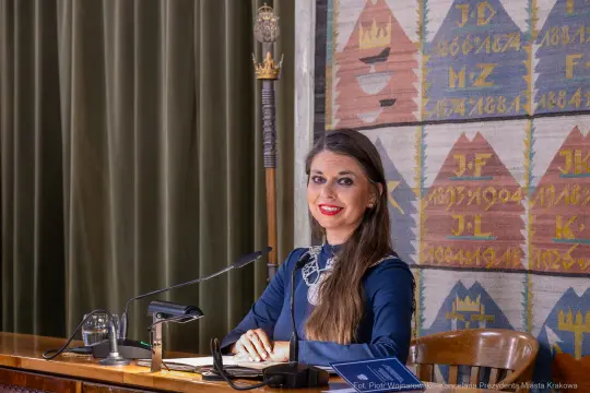 Kobieta w niebieskiej sukience siedząca przy stole, uśmiechnięta, z mikrofonem i dokumentami.