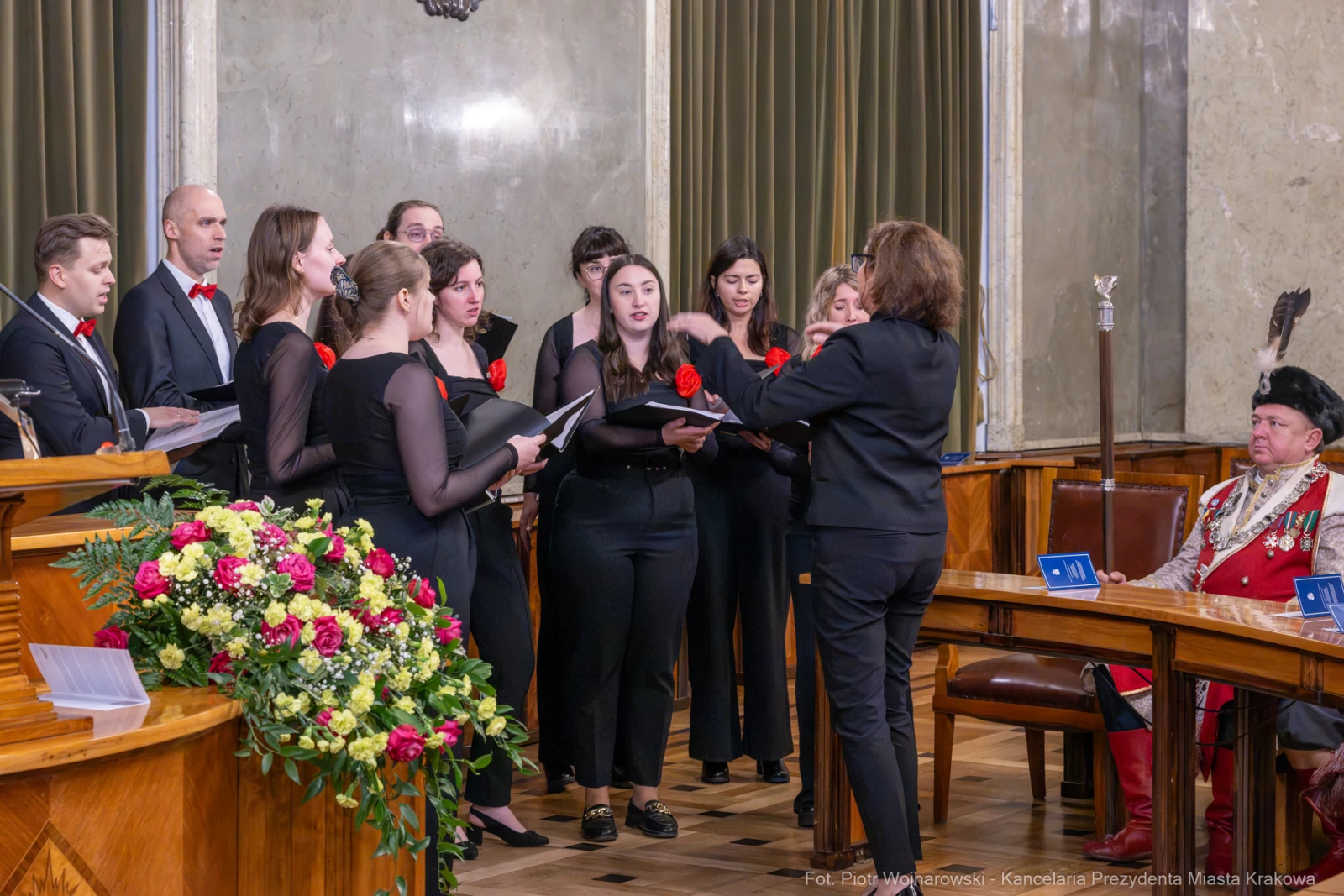 Chór w strojach formalnych wykonujący utwór w sali z dekoracjami.  Autor: Piotr Wojnarowski