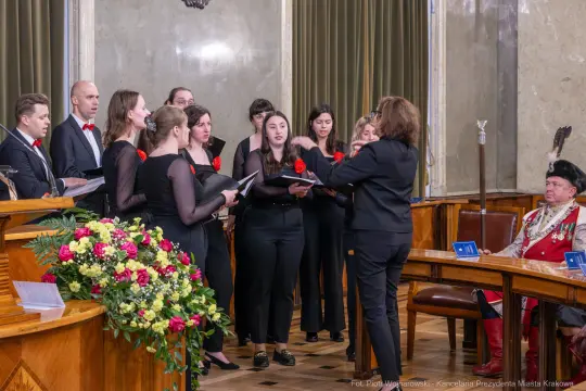 Chór w strojach formalnych wykonujący utwór w sali z dekoracjami.