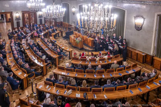 Zgromadzenie ludzi w sali obrad, występ chóru, eleganckie wnętrze z żyrandolami.