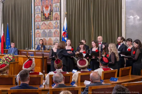Chór wykonujący utwór w sali obrad z widownią w eleganckich strojach.