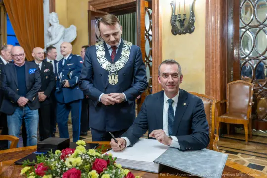 Mężczyzna podpisuje dokument w eleganckim wnętrzu, obok stoi inny mężczyzna w łańcuchu.