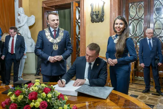 Mężczyzna podpisujący dokument w towarzystwie dwóch osób w eleganckich strojach. Kwiaty na stole.