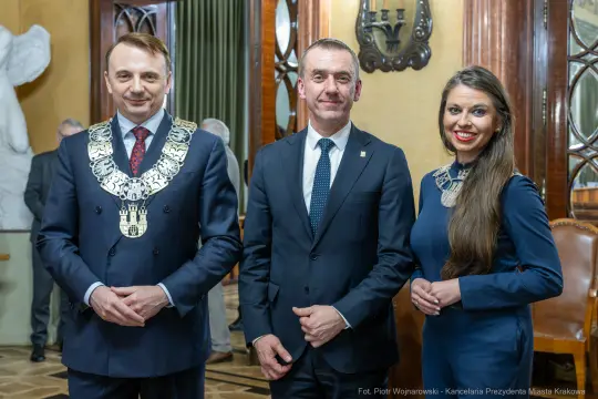 Trzech mężczyzn w garniturach i jedna kobieta w eleganckiej sukience, wszyscy z uśmiechami.