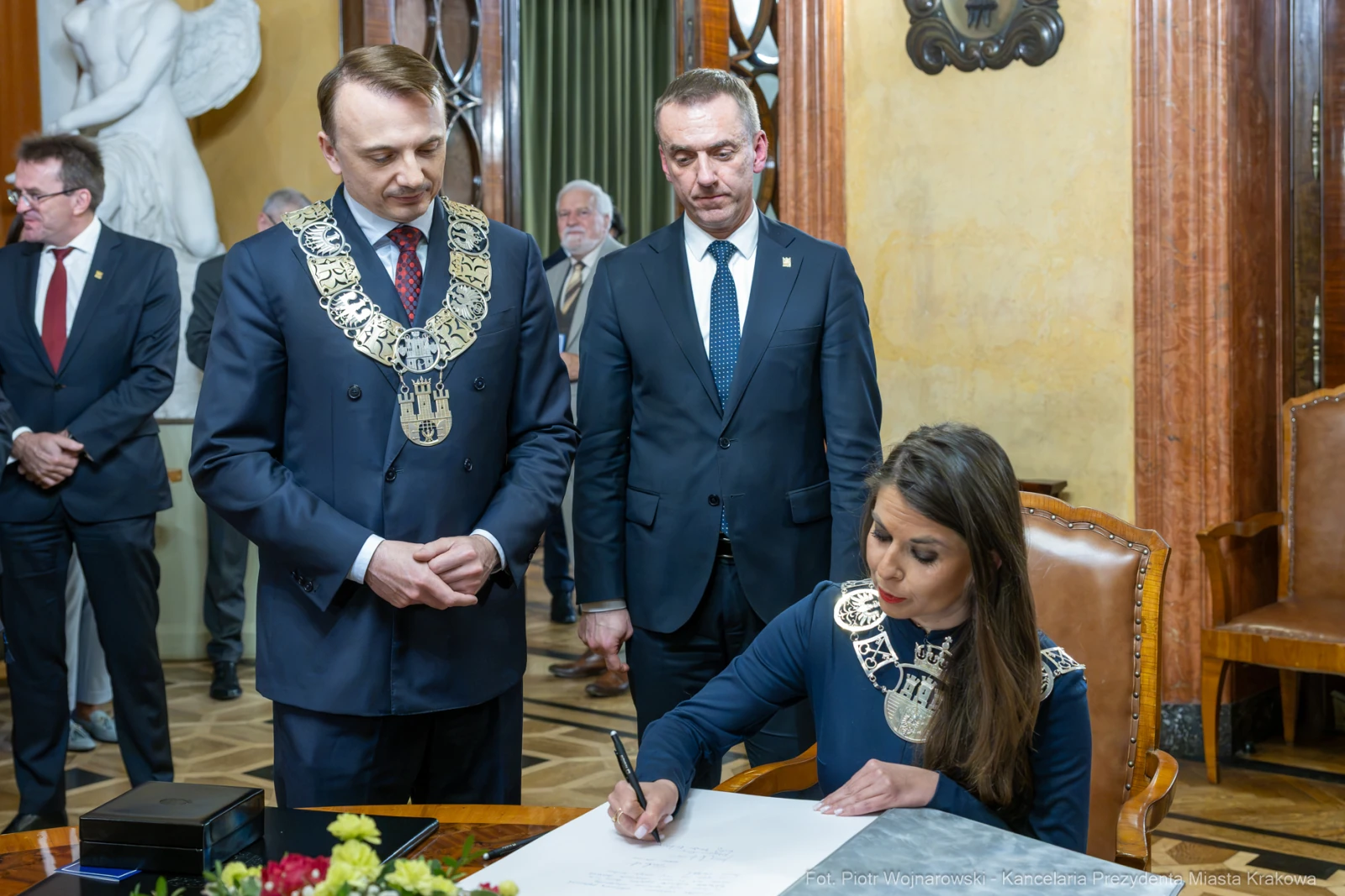 Ceremonia podpisania dokumentu przez kobietę w eleganckim stroju, mężczyźni w togach obserwują.  Autor: Piotr Wojnarowski