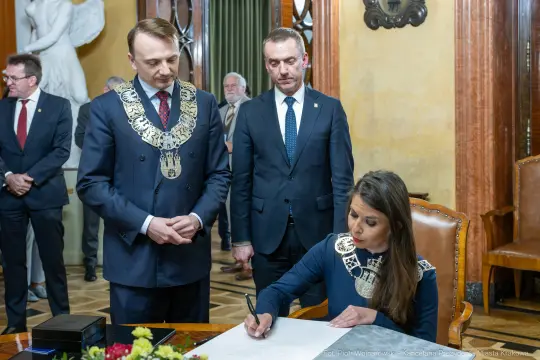 Ceremonia podpisania dokumentu przez kobietę w eleganckim stroju, mężczyźni w togach obserwują.