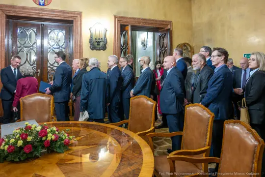 Grupa ludzi w formalnych strojach w sali z dużym stołem i dekoracjami.