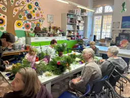 Grupa osób tworzy kompozycje florystyczne w jasnym pomieszczeniu.