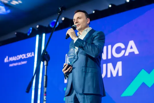Mężczyzna w niebieskim garniturze trzyma statuetkę i przemawia do mikrofonu na tle kolorowego tła.
