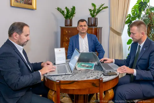 Trzech mężczyzn przy stole z laptopami, w tle komoda i dekoracje ścienne.