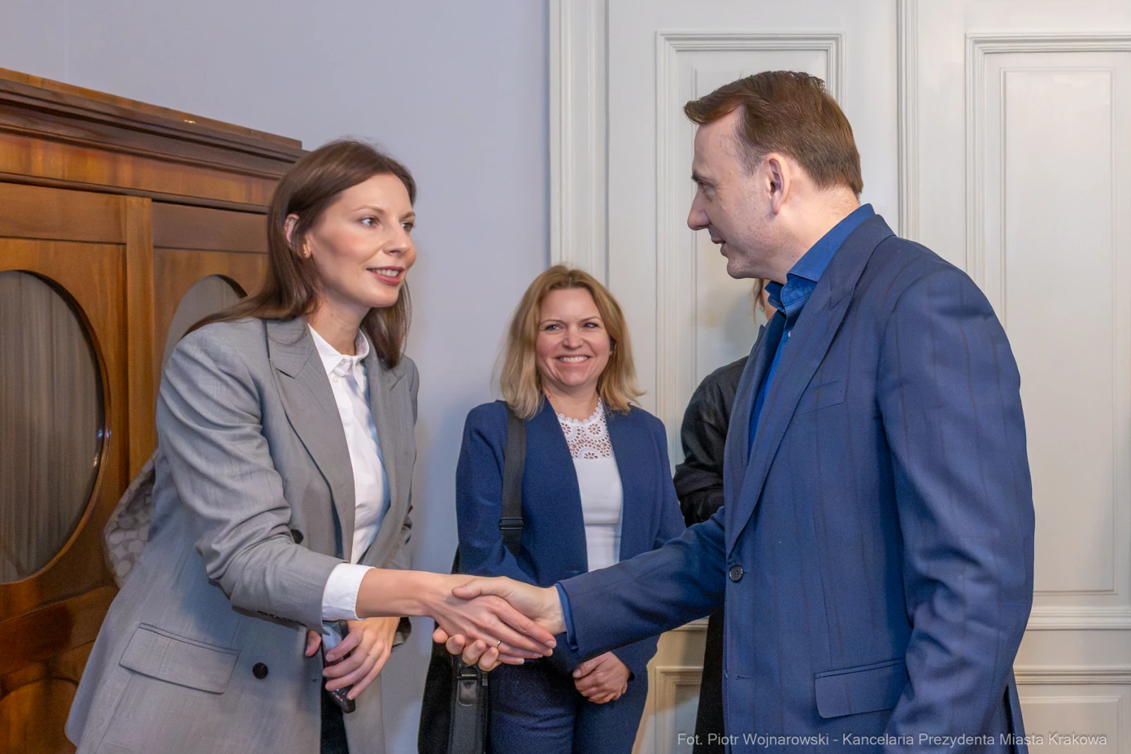 Spotkanie dwóch osób przy uścisku dłoni, w tle kobieta obserwująca sytuację.  Autor: Piotr Wojnarowski